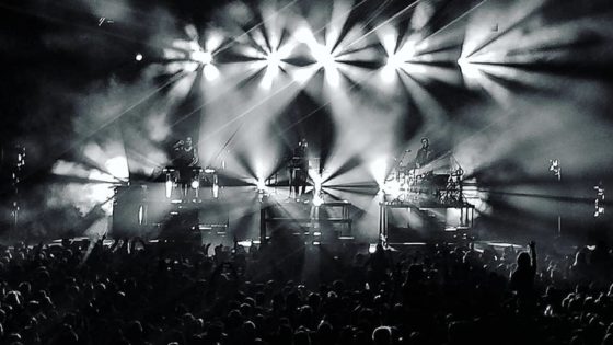 Black and white photograph depicts bright stage lights and a large crowd.