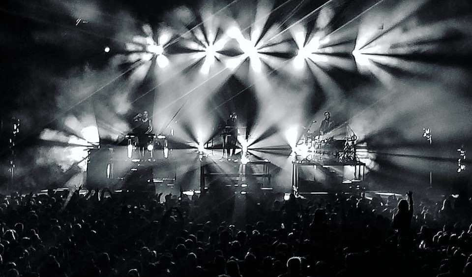 Black and white photograph depicts bright stage lights and a large crowd.