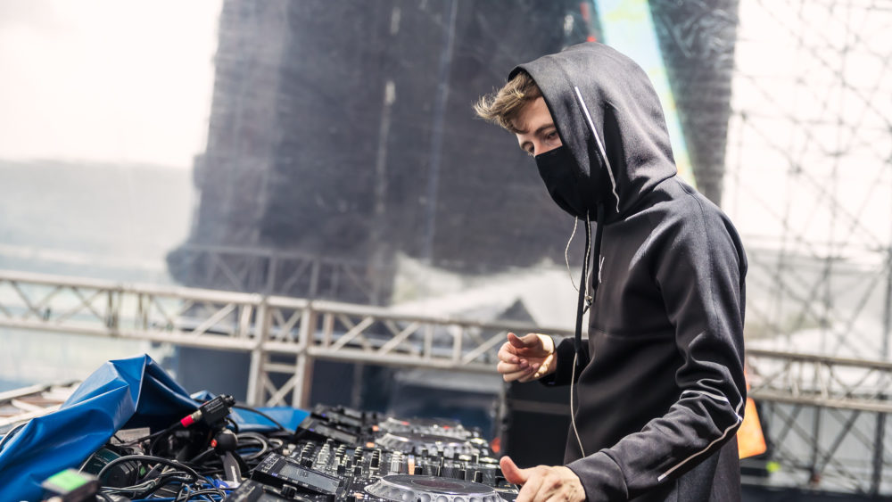 Alan Walker wearing a black hoodie and black facemask DJing at festival