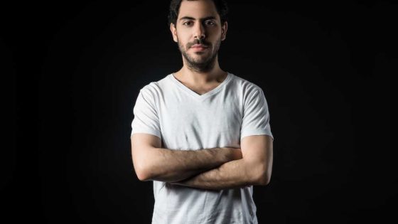 Karmy stands with arms crossed in a white shirt on black background