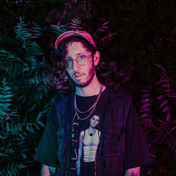 Of The Trees, standing in front of greenery. He is wearing a orange hat and a black shift with necklaces, and there is ambient soft lighting.