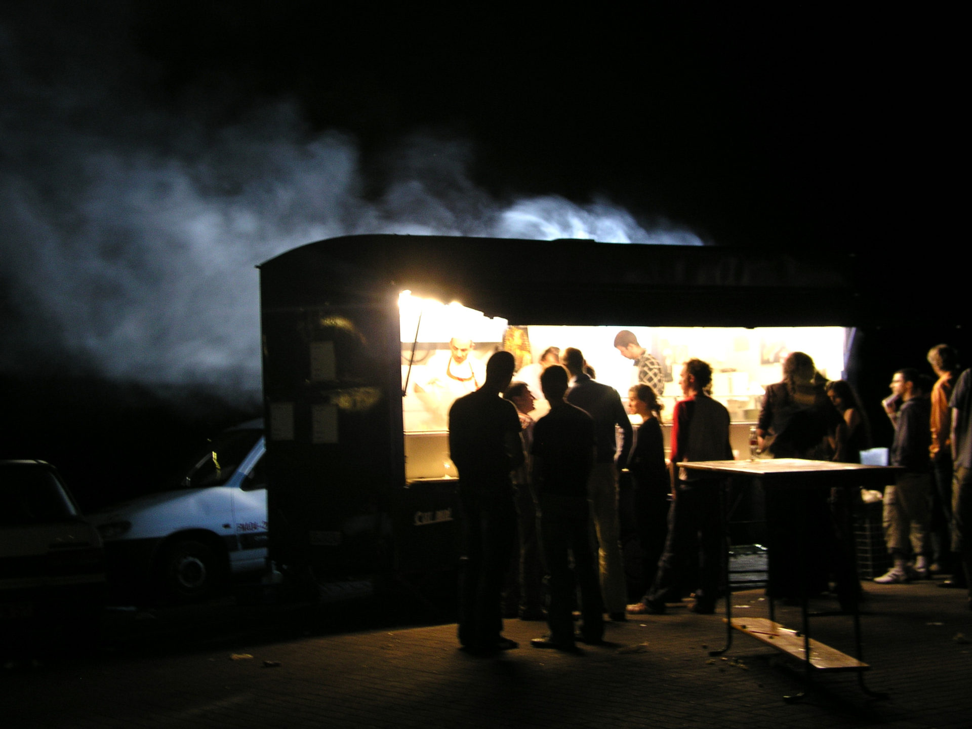 at night, a group of people is silhouetted in the light from a food cart. Smoke or steam rises in the background. 