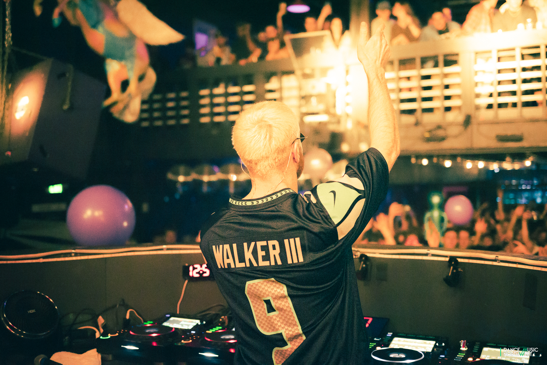 Photo from behind James Hype at DJ booth, wearing #9 Seahawks jersey in front of nightclub crowd.