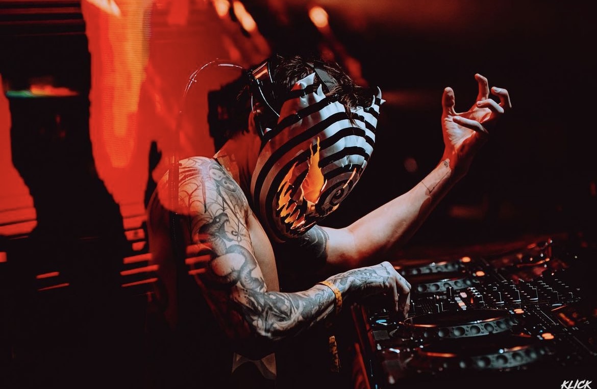 Ghastly standing on stage with his DJ board and dressed as his alternative DJ persona, Ghengar. Wearing a striped animal helmet with black and white stripes and and LED tail attached. 