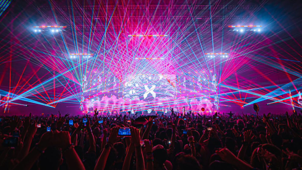 The Thunderdome stage during Excisions set. A hot pink and lavender background with purple, blue, and pink lasers shooting out into the crowd. A crowd of festival goers is seen at the bottom of the photo.