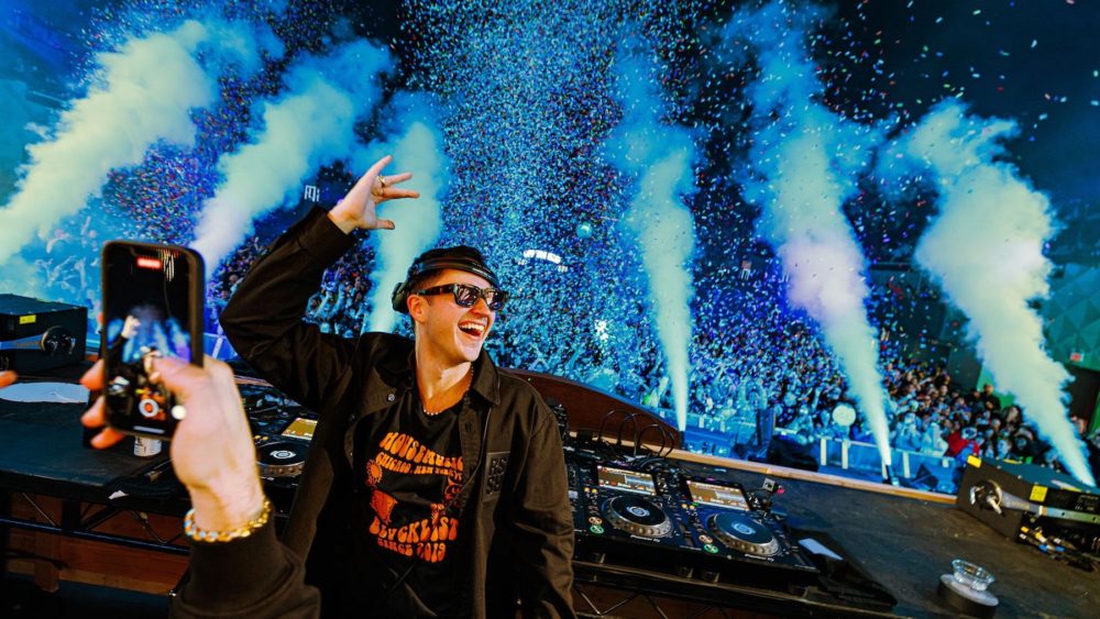 John Summit in a black jacket and black sunglasses standing in a DJ booth with confetti cannons and fog machines in blue light
