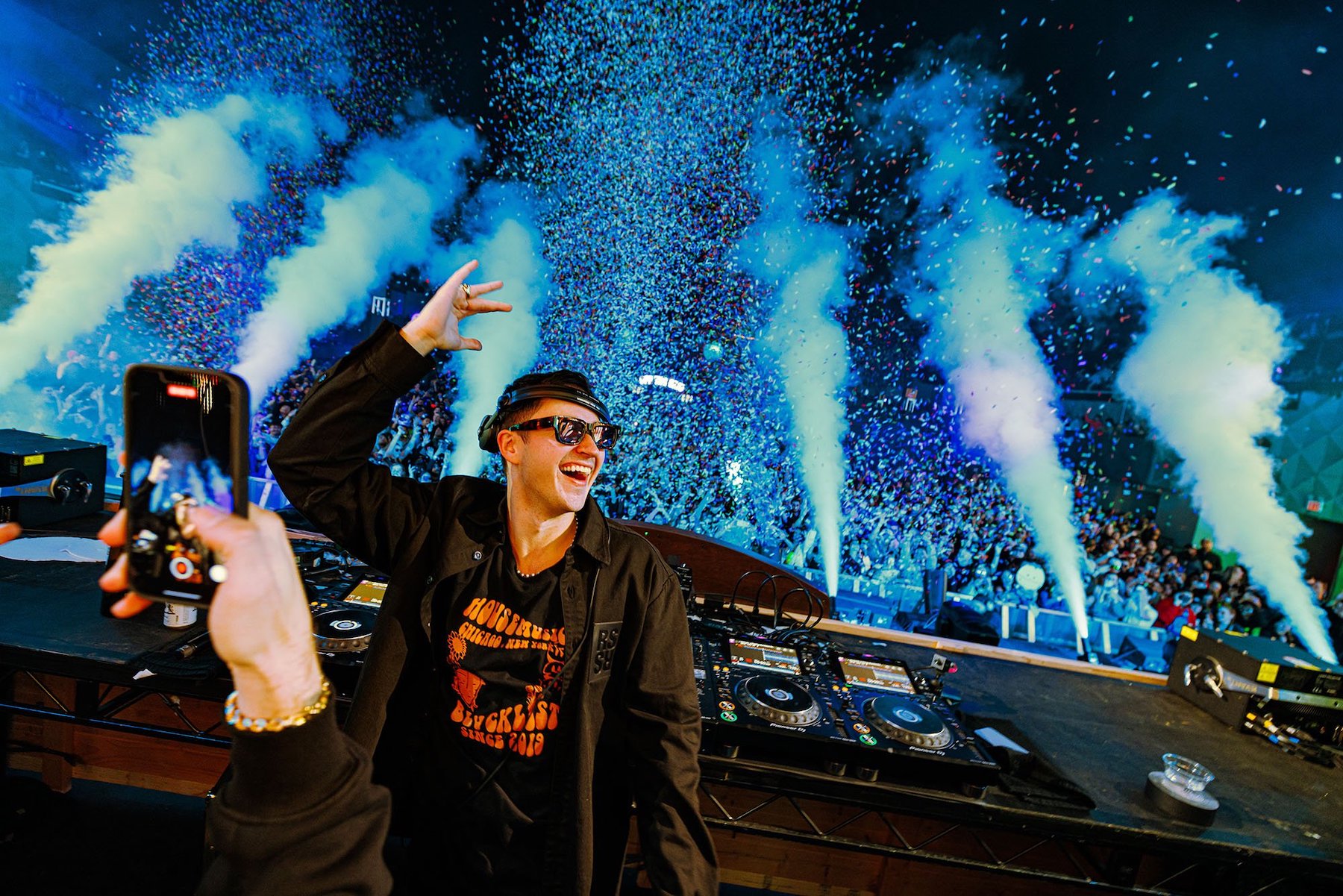 John Summit in a black jacket and black sunglasses standing in a DJ booth with confetti cannons and fog machines in blue light