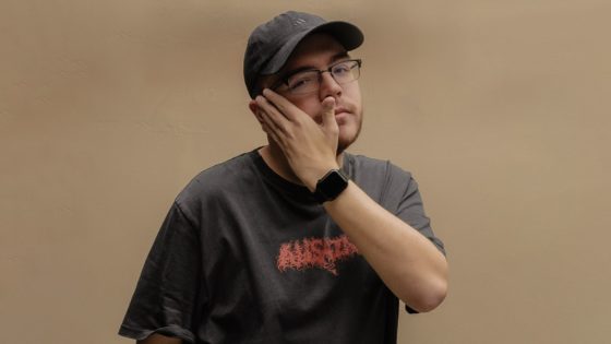 Photo of Ray Volpe from photoshoot wearing black hat and black shirt, covering right side of face with his left hand.