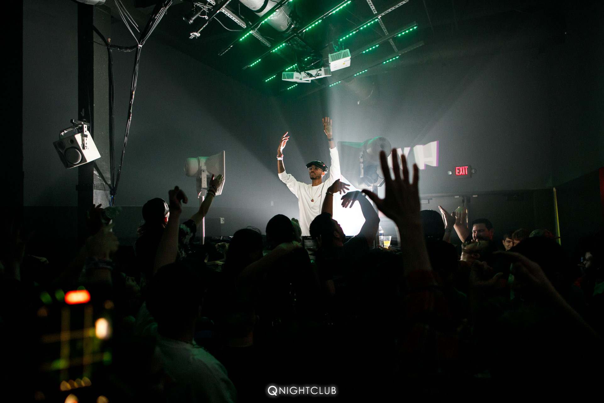 Tony H DJ'ing at Q Nightclub with hands up in air wearing white shirt, crowd also has hands in air.