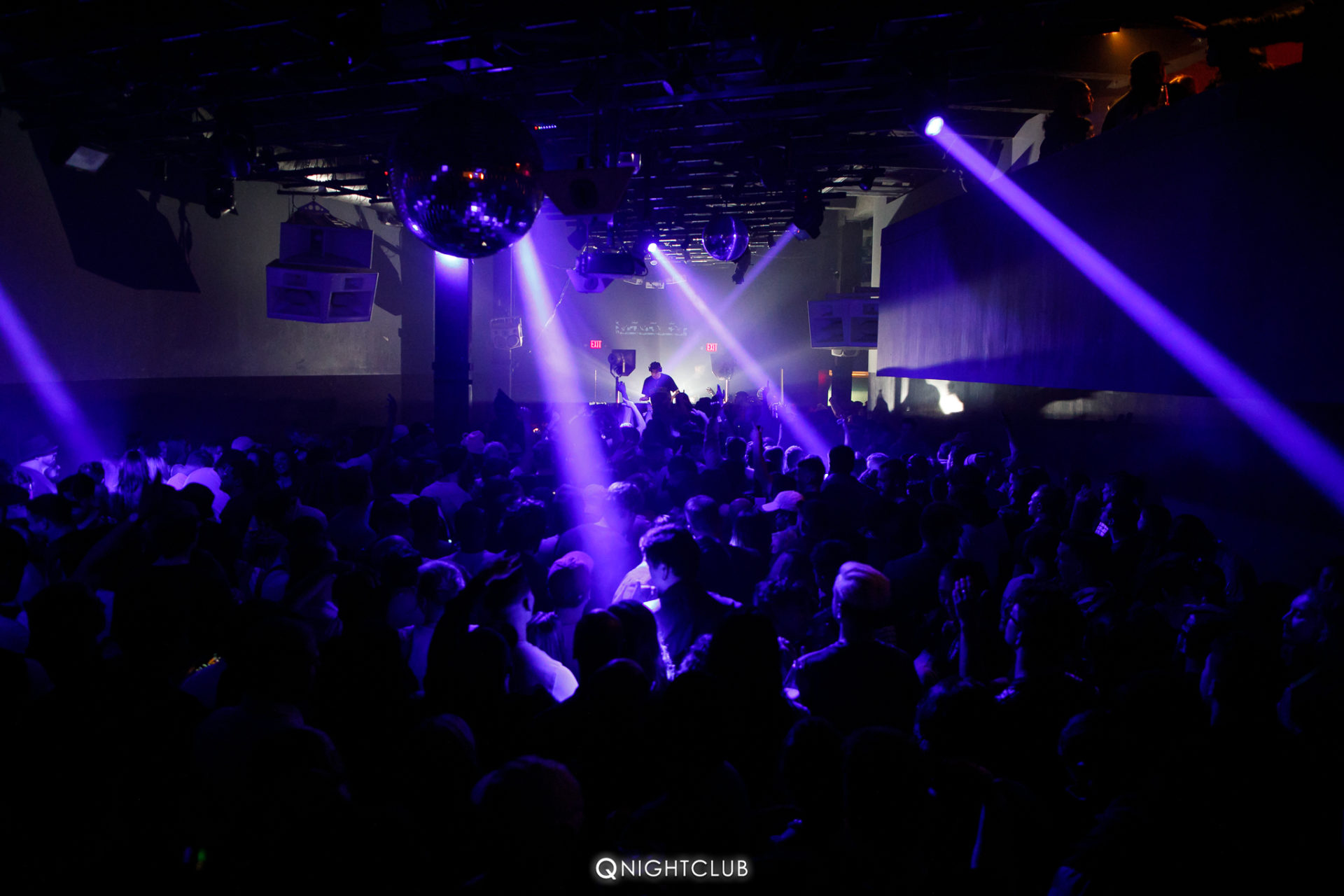 Mau P performing at Q Nightclub with purple lights from ceiling, back of the venue with crowd in front.