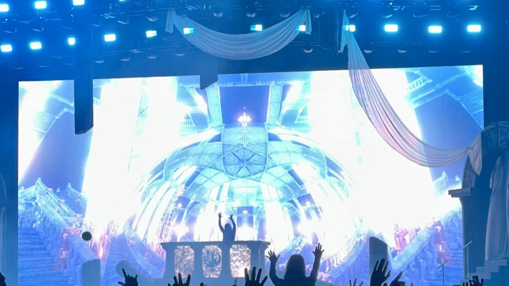 A photo of Seven Lions taken at the Tacoma Dome during the Beyond the Veil tour. Seven lions is seen Djing with his hands in the air. The background shows a blue crystal dome with blue lighting.
