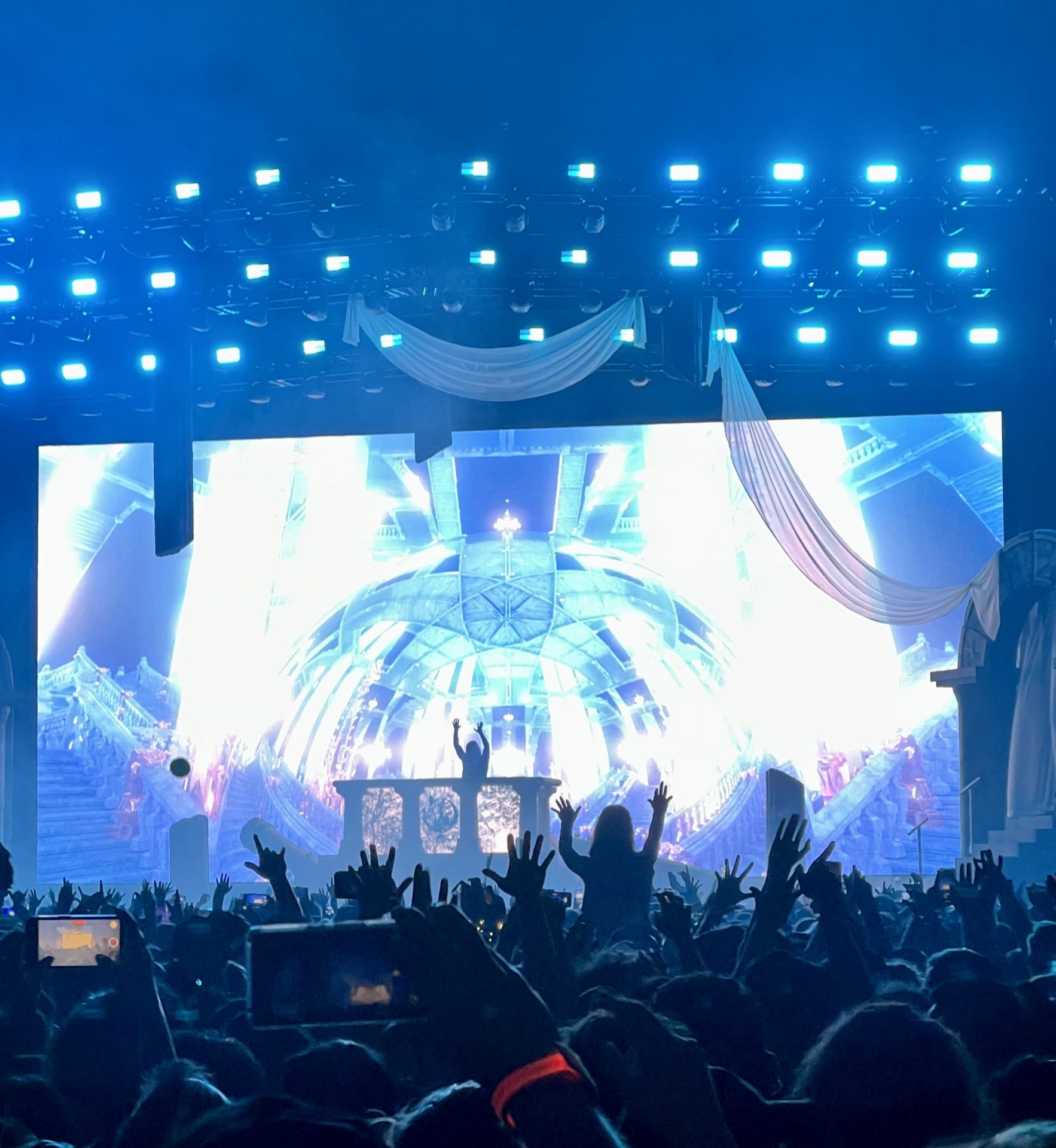A photo of Seven Lions taken at the Tacoma Dome during the Beyond the Veil tour. Seven lions is seen Djing with his hands in the air. The background shows a blue crystal dome with blue lighting.