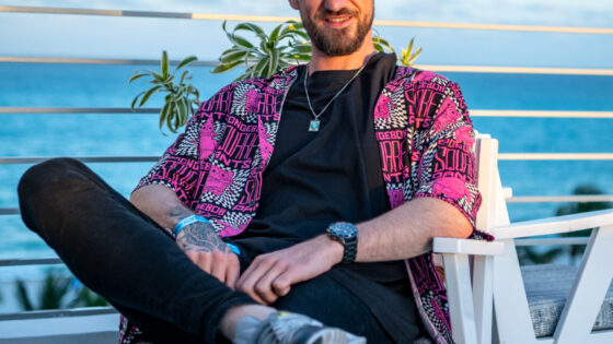 Billy Gillies sitting cross legged wearing a red shirt in front of water