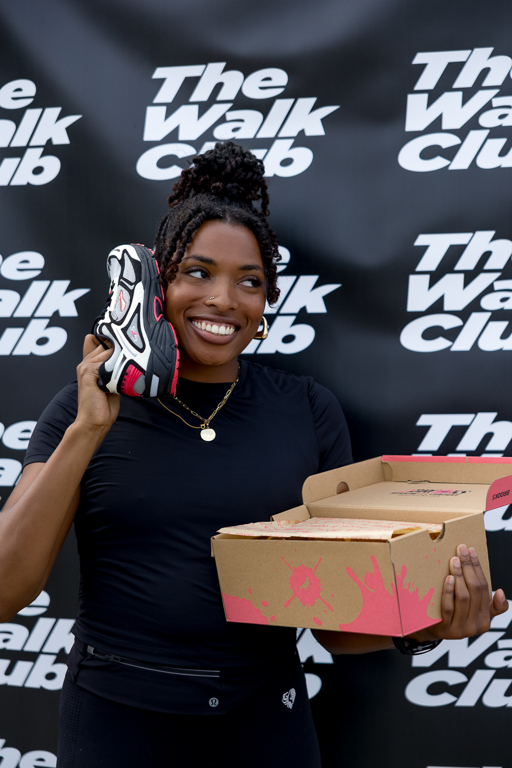 Woman holding up a Brooks Running shoe to her ear like a phone in front of a black and white The Walk Club banner