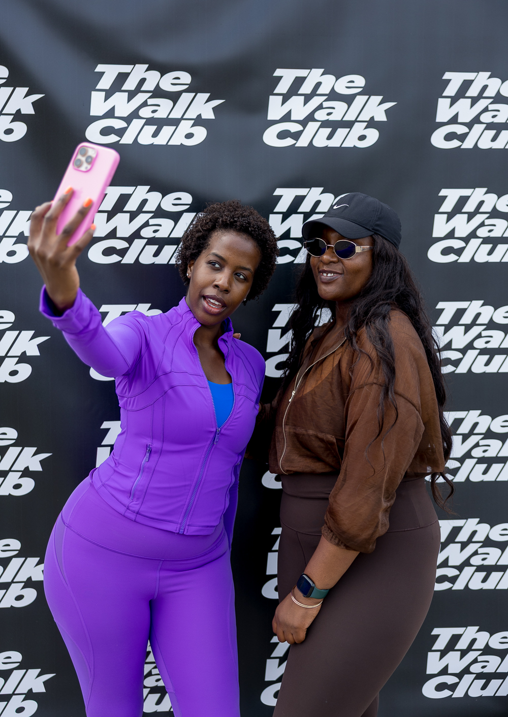 Two women taking a selfie in front of a black and white The Walk Club banner
